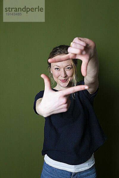 Smiling young woman in blue T-shirt gesturing finger frame in front of green wall