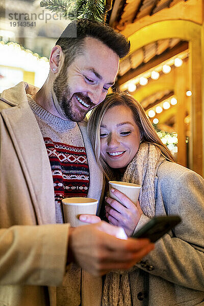 Glückliches Paar benutzt Smartphone und hält Kaffeetasse auf dem Weihnachtsmarkt