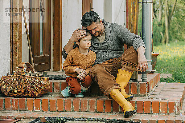 Vater umarmt Sohn  der auf den Stufen des Hauses sitzt