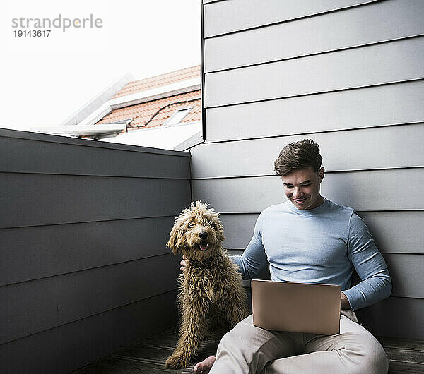 Lächelnder junger Mann benutzt Laptop und Hund sitzt auf dem Balkon