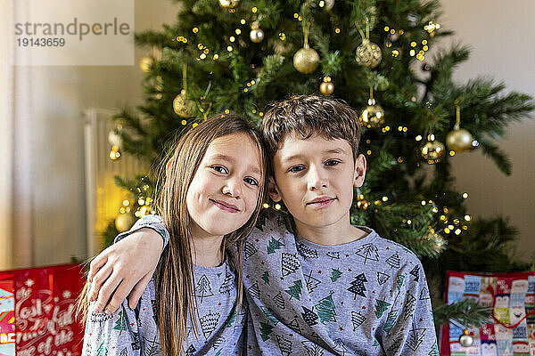 Lächelnde Schwester und Bruder sitzen zu Hause vor dem Weihnachtsbaum