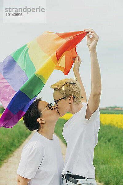 Liebevolle Lesben mit Regenbogenfahne küssen sich auf dem Feld