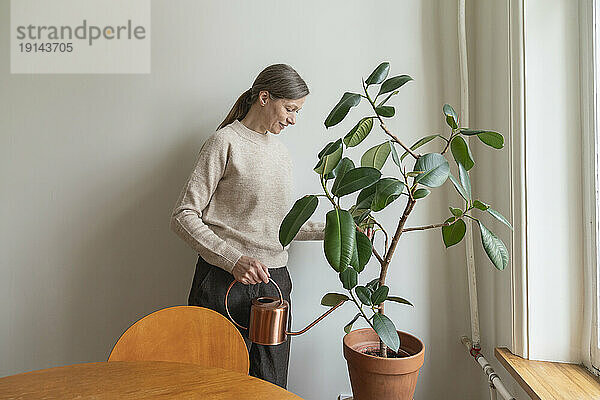 Lächelnde Frau hält Gießkanne in der Hand und kümmert sich zu Hause um Pflanzen