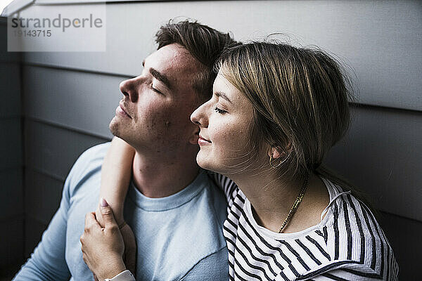 Lächelnde Frau und Mann sitzen mit geschlossenen Augen an der Wand auf dem Balkon