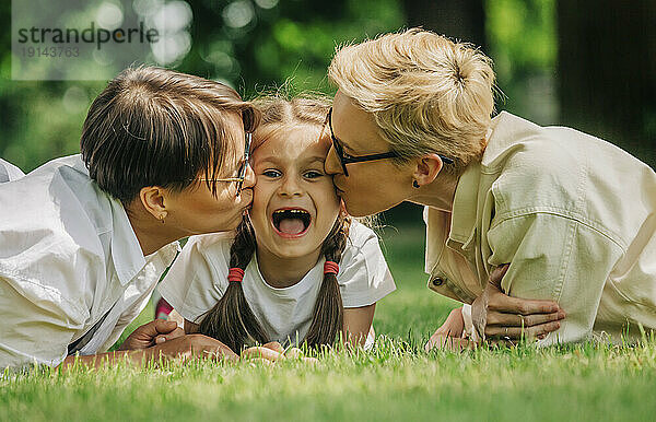 Lesbian couple kissing daughter lying on grass at park