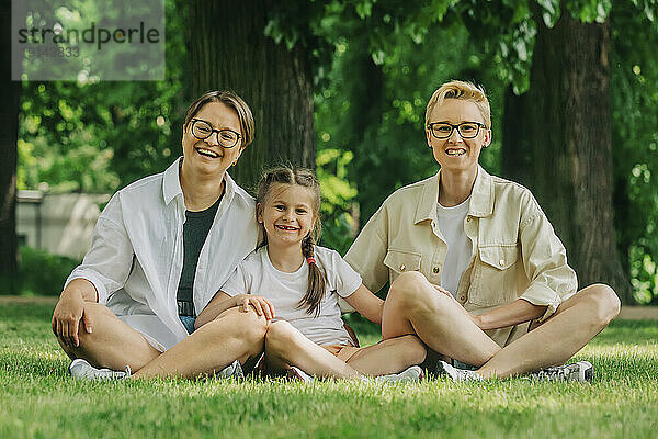 Smiling lesbian couple with daughter sitting cross-legged at park