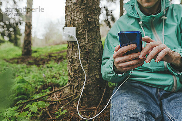 Mann benutzt Smartphone mit Steckdose am Baumstamm