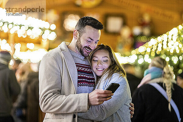 Glücklicher Mann und Frau  die auf dem Weihnachtsmarkt ihr Mobiltelefon benutzen