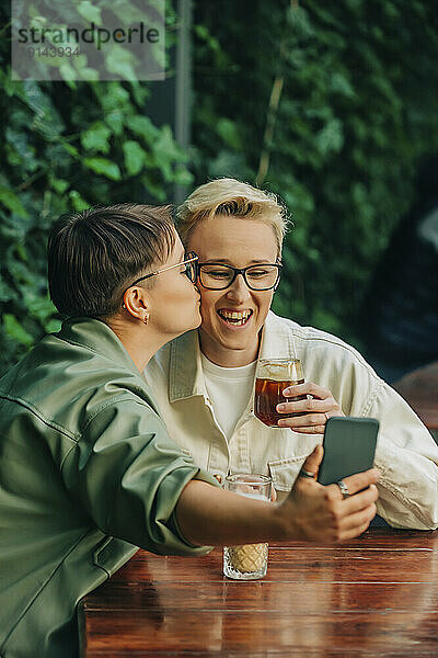 Smiling lesbian woman kissing on cheek of girlfriend and taking selfie through smart phone