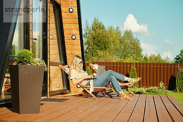 Frau sieht sich einen Film auf dem Laptop an  während sie sich in einem klappbaren hölzernen Liegestuhl auf der Terrasse einer Blockhütte ausruht