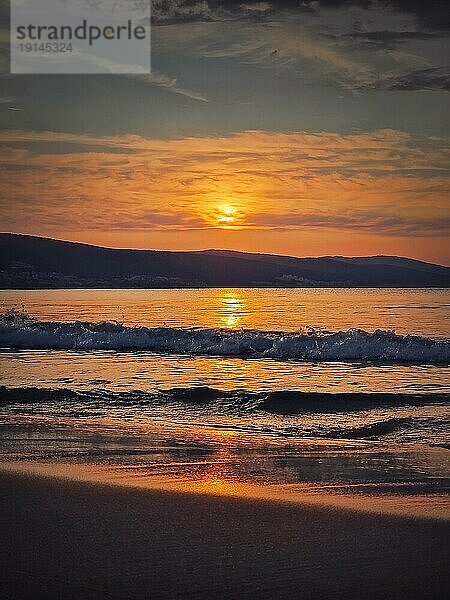 Frühmorgens am Strand mit einem leuchtenden Sonnenaufgang am Horizont und schäumenden Wellen  die auf den Sand schlagen. Morgendämmerung am Meer  friedliche Aussicht