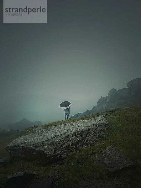 Rückansicht eines einsamen Mannes mit Regenschirm steht auf einem felsigen Hügel  der von Dunst bedeckt ist. Stimmungsvolle und emotionale Szene mit einem einsamen Fremden Silhouette unter dem regen