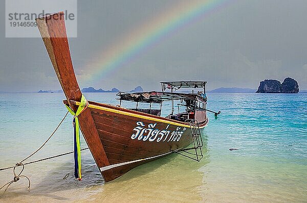 Longtailboot in kristallklarem blaün Wasser wartet auf Passagiere auf schönen weißen Strand in Ko Mok  Thailand nach regen mit einem schönen Regenbogen