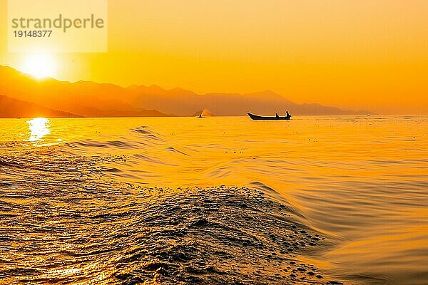 Silhouette eines Fischerboots mit einem Fischer beim Fischen im orangefarbenen Sonnenuntergang des Shkoder Sees in Shiroka und den Bergen. Albanien