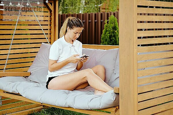 Mittlere erwachsene Frau entspannt sich auf einer Schaukel im Garten und schaut auf ihr Smartphone