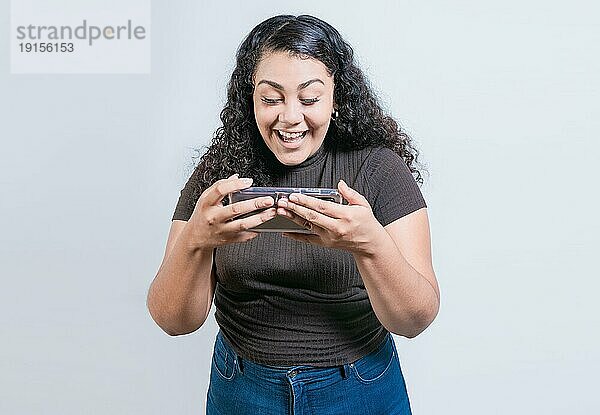 Glückliches lateinisches Mädchen  das ein Mobiltelefon auf einem isolierten Hintergrund benutzt. Fröhliche Menschen schauen auf eine Förderung auf dem Telefon isoliert