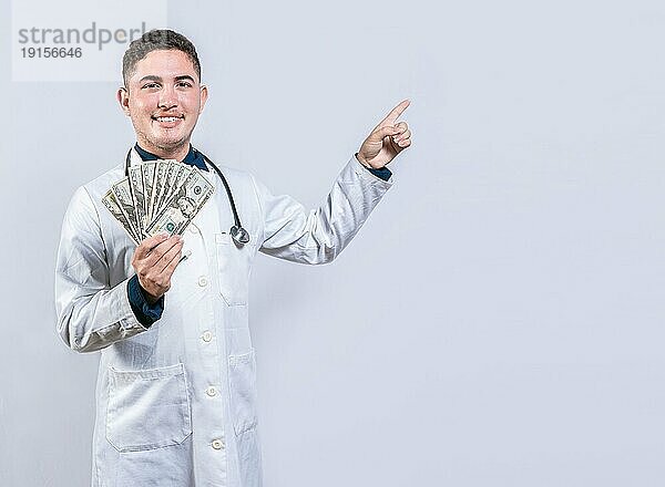 Glücklicher lateinischer Arzt  der Geld hält und auf die Seite zeigt. Hübscher lächelnder Arzt zeigt Geld und zeigt auf die Seite