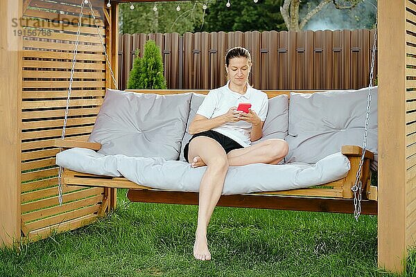 Erwachsene Frau genießt friedlichen Urlaub ruht mit Smartphone in bequemen Schaukel auf Hinterhof