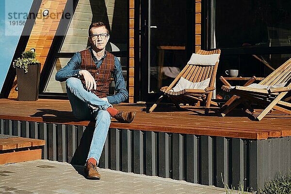 Mann mittleren Alters in Jeans  Hemd und Weste sitzt morgens auf der Terrasse einer Holzhütte