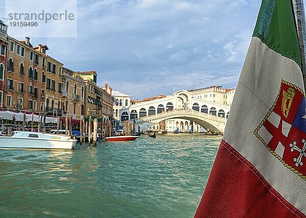 Schöner Blick auf die italienische Seeflagge und die Rialtobrücke oder Ponte Rialto am Canal Grande in Venedig  Italien. Gondel Service Station auf der linken Seite. UNESCO Weltkulturerbe Stadt. Tageslicht  Sonnenschein  weiche Wolken. Selektiver Fokus