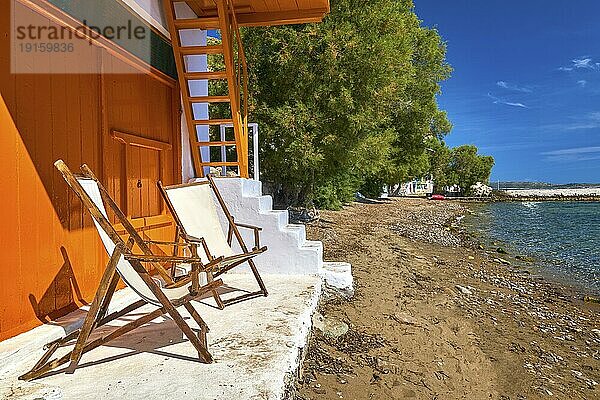 Zwei hölzerne Strandstühle auf der Veranda eines traditionellen griechischen Fischerhauses direkt am Meer. Weiß getünchte Hauswände und orange gestrichene Türen und Treppen  keine Menschen  Sommersonne  sonniger Tag  Entspannung  meditativer Moment  perfekter Urlaubsort