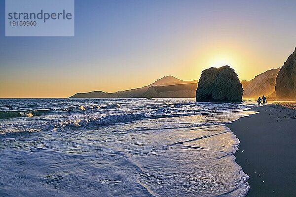 Entfernte Silhouetten von zwei Erwachsenen zu Fuß in den Gezeiten am Strand von Fyriplaka  Insel Milos  Kykladen  Griechenland. Schöne Sonnenuntergang Himmel  einzigartige einsamen Felsen am Sandstrand  niedrige Sonne  Sonnenuntergang Dunst  felsigen Hügeln