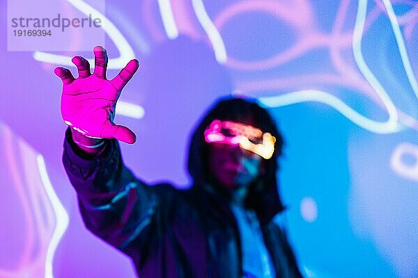 Studioporträt mit lila und blauen Neonröhren mit Fokus auf die Hand eines futuristischen Mannes mit AR Brille