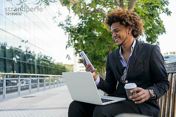 Cooler afroamerikanischer Geschäftsmann mit Laptop und Handy auf der Straße sitzend am Morgen