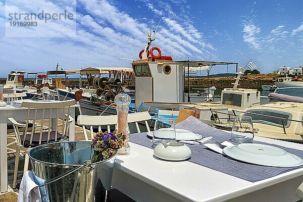 Tische und Stühle eines Fischrestaurants am Ufer der Bucht von Naoussa  neben den Tischen vertäute Fischerboote in der Sonne des Sommertages. Traditionelle griechische Küche  Urlaub  Reiseziel  Touristenparadies  romantisches Essen  selektiver Fokus