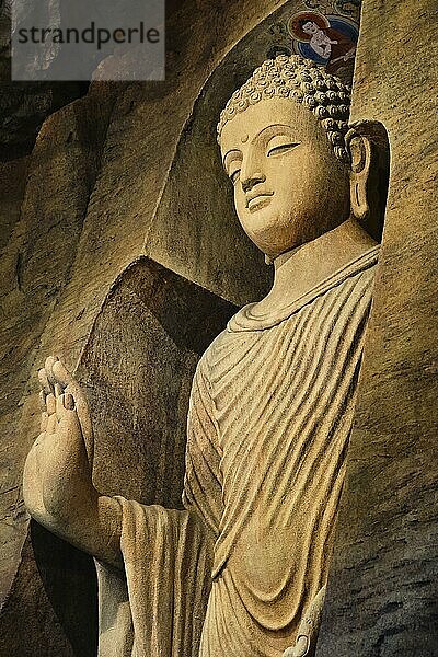 Stehende Buddhastatue in Mönchskleidung  die in eine Kalksteinhöhle gemeißelt und von Licht beleuchtet ist. Rechte Hand in Adhay Mudra Geste. Religion  Tradition  Gebet  Meditation