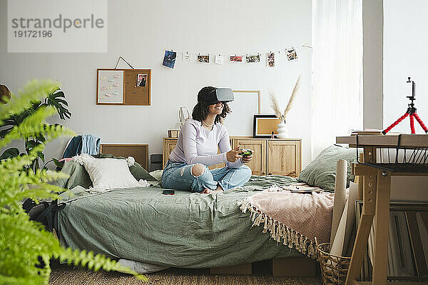 Smiling girl wearing virtual reality simulators holding game controller on bed at home