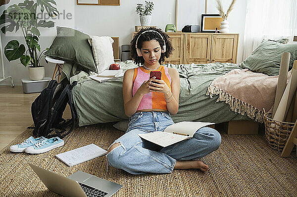 Teenager-Mädchen trägt kabellose Kopfhörer und benutzt ihr Mobiltelefon auf dem Teppich zu Hause