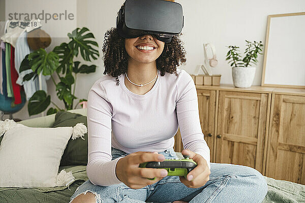 Happy girl wearing virtual reality simulators holding game controller on bed at home