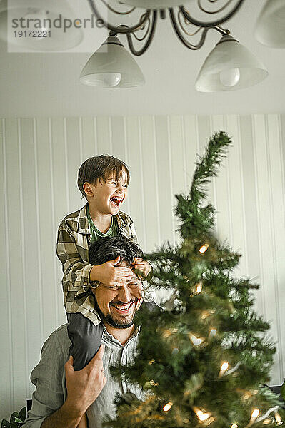 Glücklicher Vater trägt seinen Sohn über der Schulter neben dem Weihnachtsbaum zu Hause