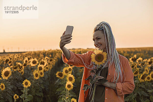 Lächelnde Frau macht ein Selfie mit ihrem Smartphone und hält eine Sonnenblume im Feld