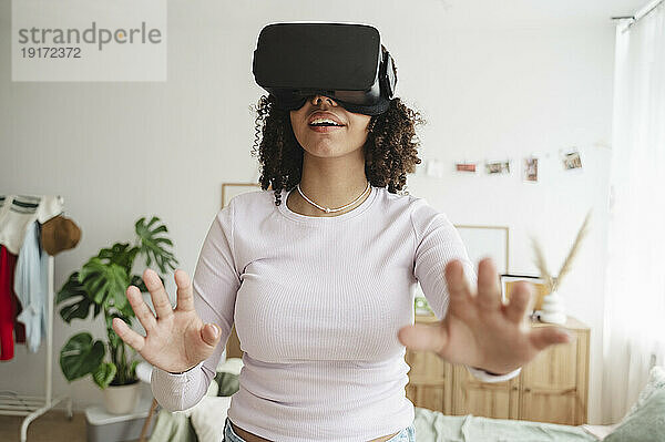 Fröhliches Teenager-Mädchen mit Virtual-Reality-Simulatoren gestikuliert im heimischen Schlafzimmer