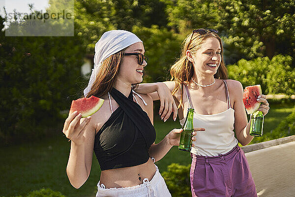 Glückliche Frau mit Freundin  die an einem sonnigen Tag Flasche und Wassermelone hält
