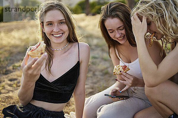 Lächelnde junge Frau mit Pizzastück von Freunden im Park