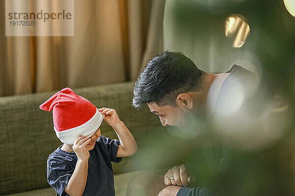 Verspielter Sohn bedeckt Gesicht mit Weihnachtsmütze auf Sofa zu Hause