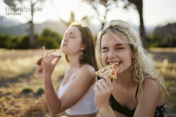Junge Frau zwinkert und isst Pizza mit einer Freundin im Hintergrund