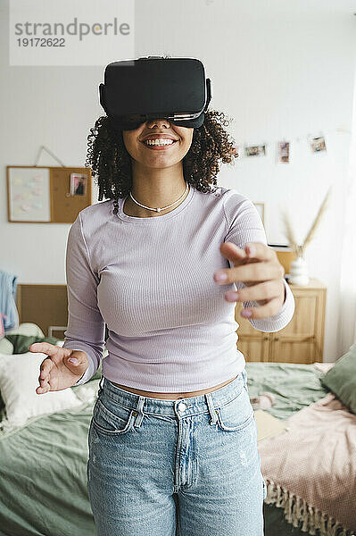 Fröhliches Mädchen mit Virtual-Reality-Simulatoren gestikuliert im Schlafzimmer