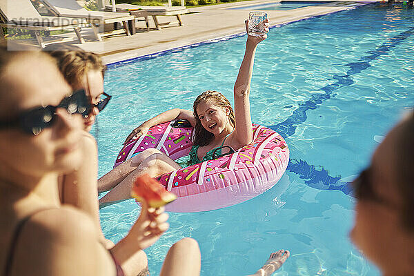 Fröhliches Teenager-Mädchen mit erhobenem Arm  Glas in der Hand und im Pool sitzend