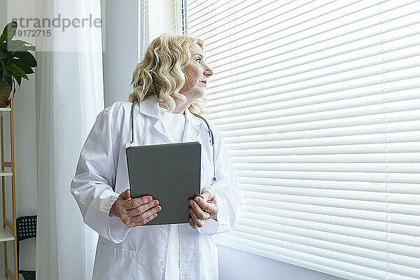 Nachdenklicher Oberarzt mit Tablet-PC  der durch das Fenster schaut