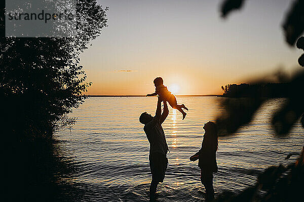 Mutter und Vater halten ihren Sohn bei Sonnenuntergang vor dem Meer