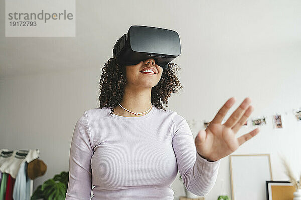Smiling curly haired girl wearing virtual reality simulators gesturing at home