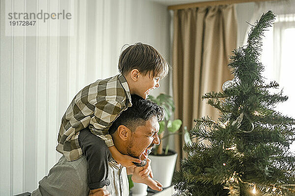 Vater trägt Sohn über der Schulter und lacht zu Hause neben dem Weihnachtsbaum