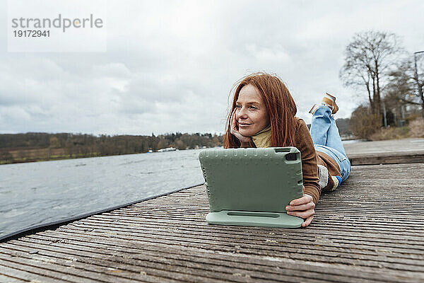 Nachdenkliche Frau mit Tablet-PC liegt am Pier am See