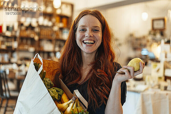 Glückliche rothaarige Frau hält Birnenfrüchte in der Hand und erledigt Lebensmitteleinkäufe