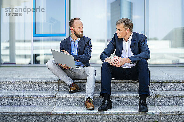 Zwei Geschäftskollegen sitzen auf Stufen und arbeiten gemeinsam am Laptop