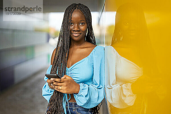 Lächelnde junge Frau steht mit Smartphone in der Nähe der gelben Wand